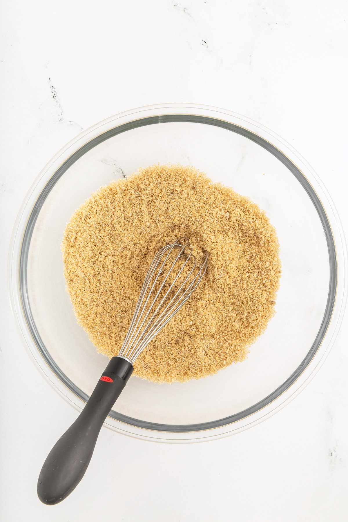 A glass bowl filled with brown sugar and a metal whisk with a black handle resting inside on a white surface.