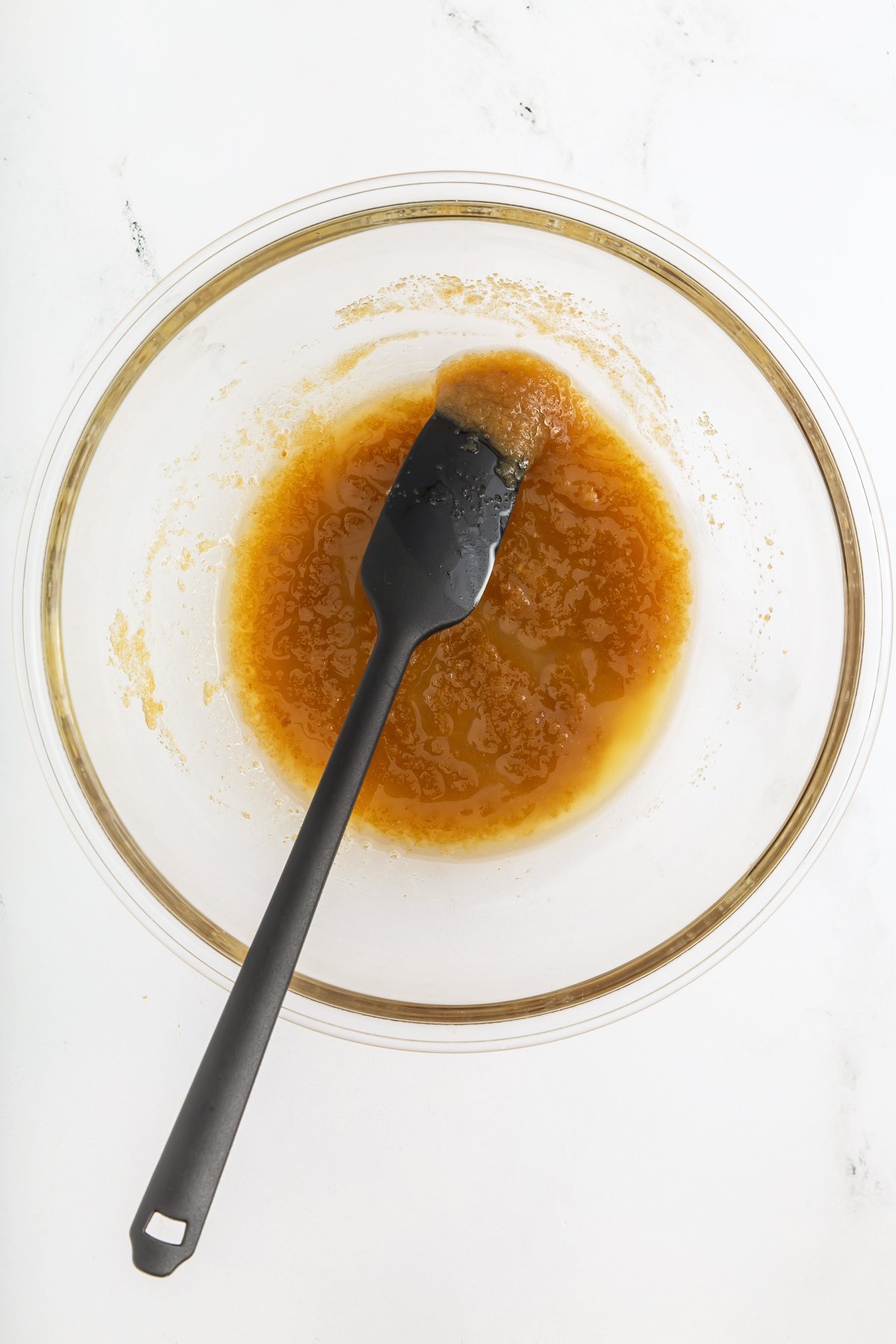 A glass bowl with a light brown liquid mixture and a black spatula resting inside, on a white surface.