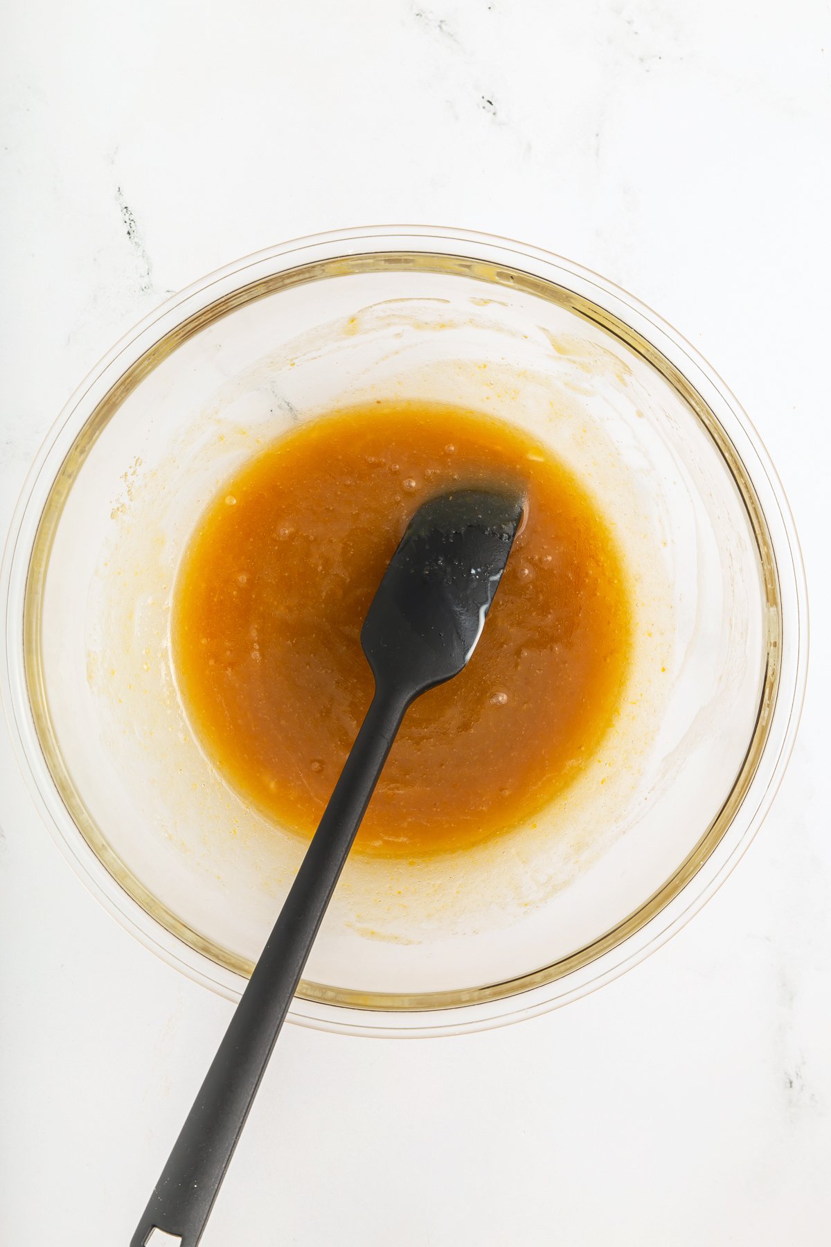 A glass bowl with a thick orange-brown liquid mixture and a black spatula resting inside, on a white surface.