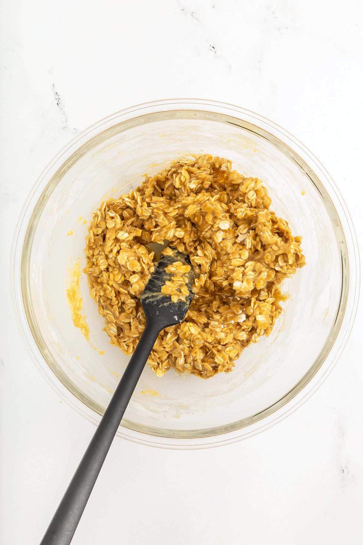 A glass bowl containing a mixture of oats and a yellow batter, being stirred with a black spoon, on a white surface.