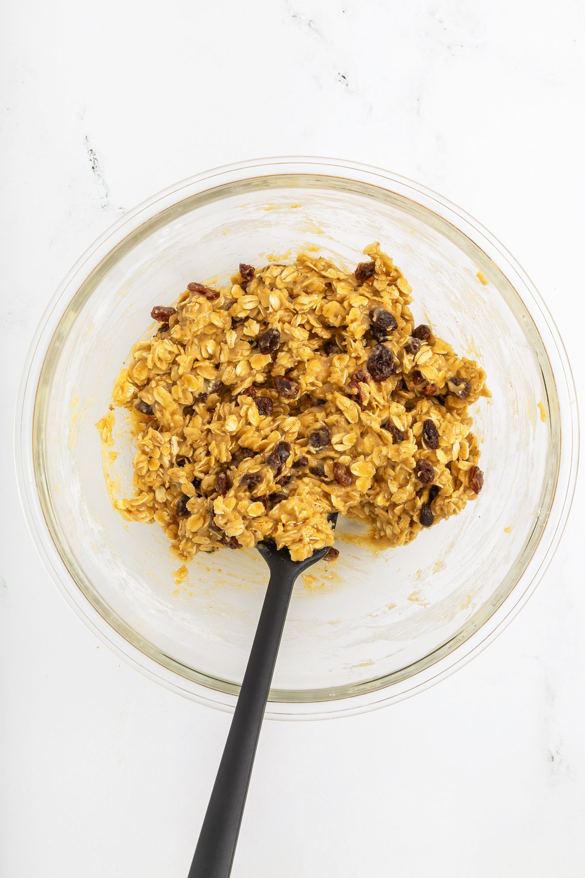 A glass bowl containing an oatmeal mixture with raisins, being stirred with a black spatula on a white surface.