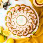 A bundt cake with white icing is displayed on a plate, surrounded by fresh lemons and a yellow napkin.
