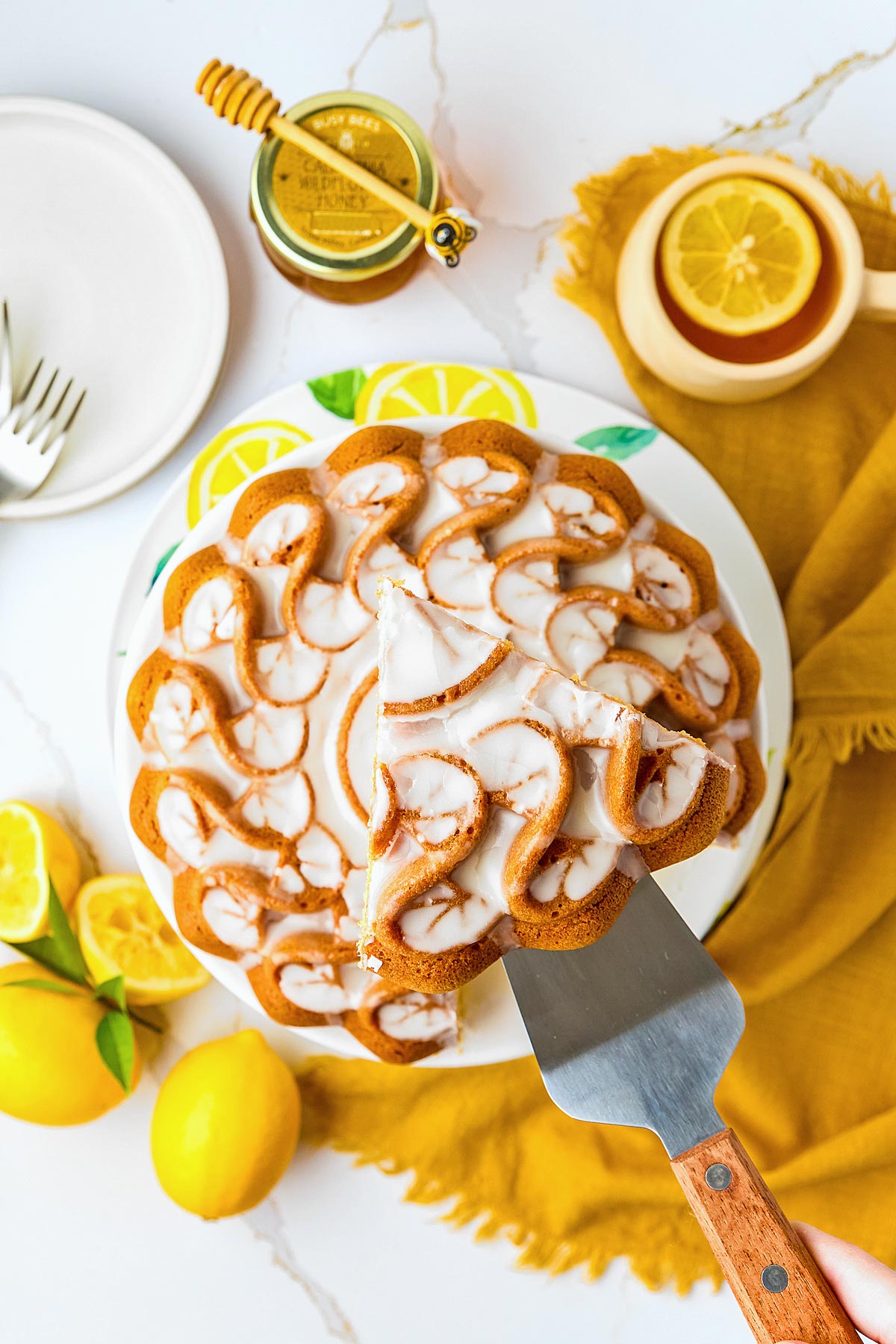 Overhead view of a lemon bundt cake with white glaze, one slice being served, surrounded by fresh lemons, honey, and a cup with a lemon slice.