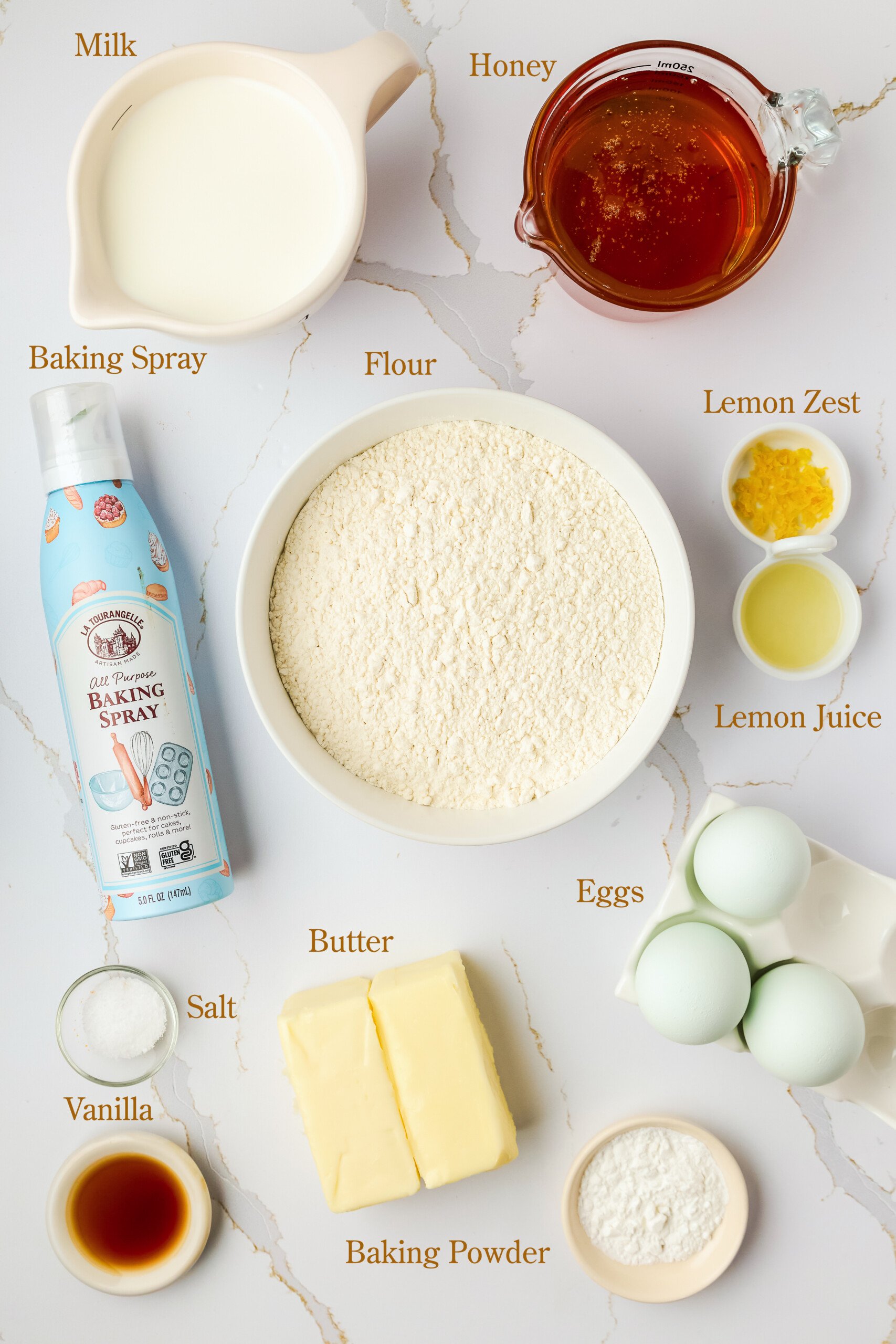 Various baking ingredients are arranged on a white surface, including milk, honey, flour, lemon zest, lemon juice, eggs, butter, baking spray, vanilla, salt, and baking powder.