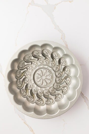 A round, silver Bundt cake pan with an intricate swirl and floral pattern sits on a white marble surface with light brown veining.