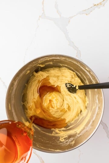 A metal mixing bowl with creamed butter and sugar, with liquid honey being poured in, and a black spatula resting inside the bowl.