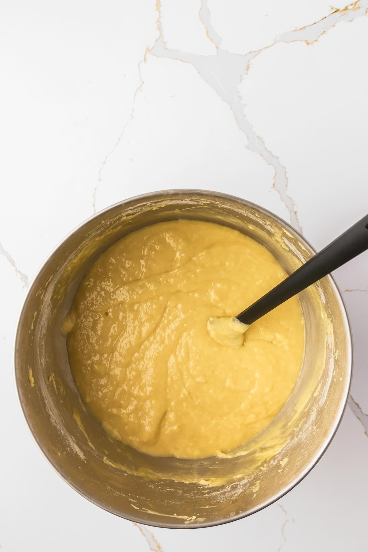 A metal bowl filled with yellow cake batter and a black spatula resting inside, placed on a white marble countertop.