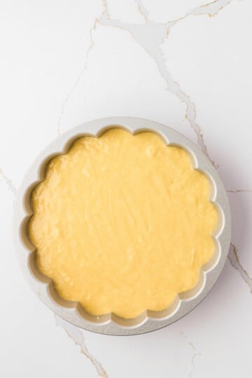 A round scalloped-edge cake pan filled with raw yellow cake batter, placed on a white marble surface.