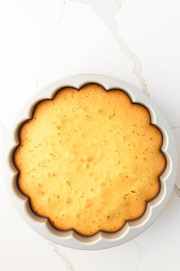 A golden-brown round cake with scalloped edges in a silver baking pan, viewed from above on a white marble surface.