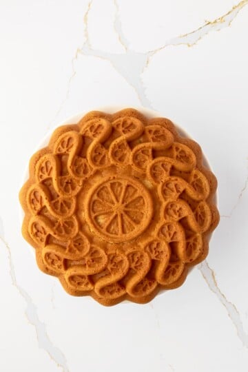 A round, golden-brown bundt cake with an intricate floral pattern sits on a white surface with subtle gold veining.