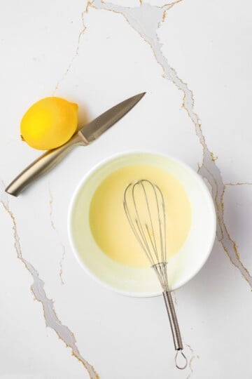 A lemon, a knife, and a bowl of pale yellow mixture with a whisk are arranged on a white marble surface.