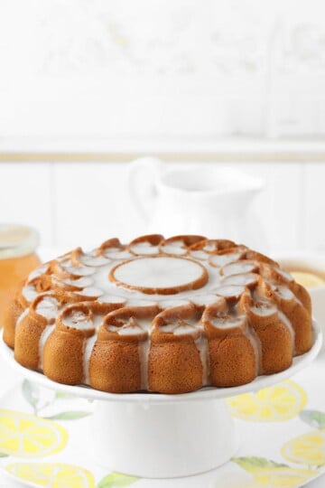 A round bundt cake with white icing drizzled on top, displayed on a white cake stand in a bright kitchen setting.