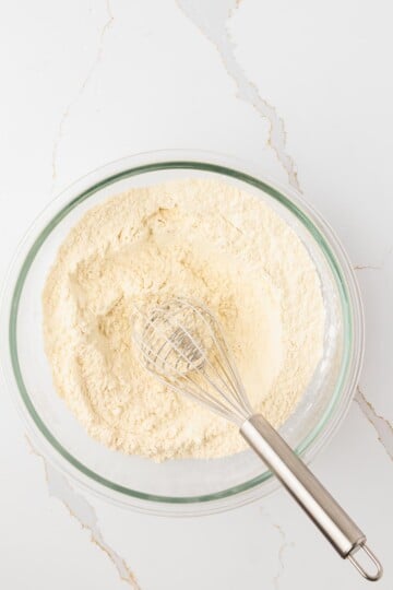 A glass bowl filled with flour mixture and a metal whisk resting inside, placed on a white countertop.
