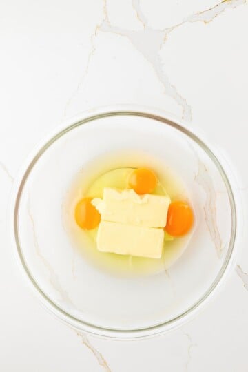 A glass bowl containing two eggs and two sticks of butter on a white marble surface.