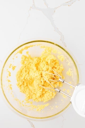 A glass bowl contains a yellow dough mixture being mixed with an electric hand mixer on a white marble surface.