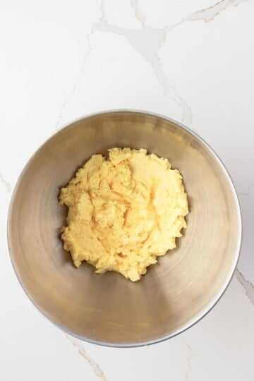 A stainless steel mixing bowl containing a yellow, creamy mixture on a white marble surface.