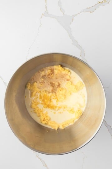 A metal mixing bowl containing milk, eggs, and vanilla extract on a white marble countertop.