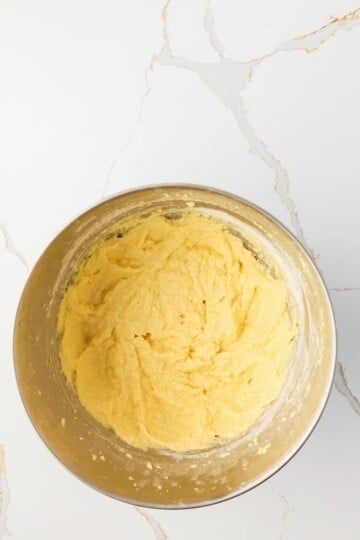 A metal mixing bowl filled with yellow cake batter sits on a white marble countertop.