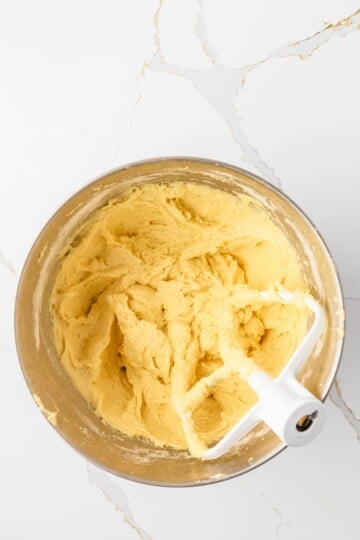 A metal mixing bowl containing yellow cookie dough and a white paddle attachment sits on a white marble countertop.