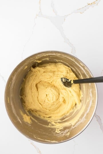 A stainless steel bowl containing yellow cake batter being mixed with a black spatula, placed on a white marble countertop.