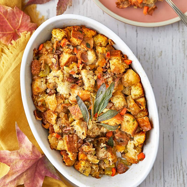 Oval white dish filled with bread stuffing containing vegetables and herbs, garnished with sage leaves, set on a light table with autumn leaves.