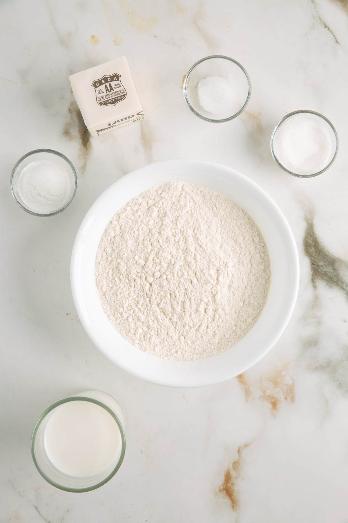 A bowl of flour, a glass of milk, a stick of butter, and three small bowls containing salt, sugar, and yeast arranged on a marble surface.