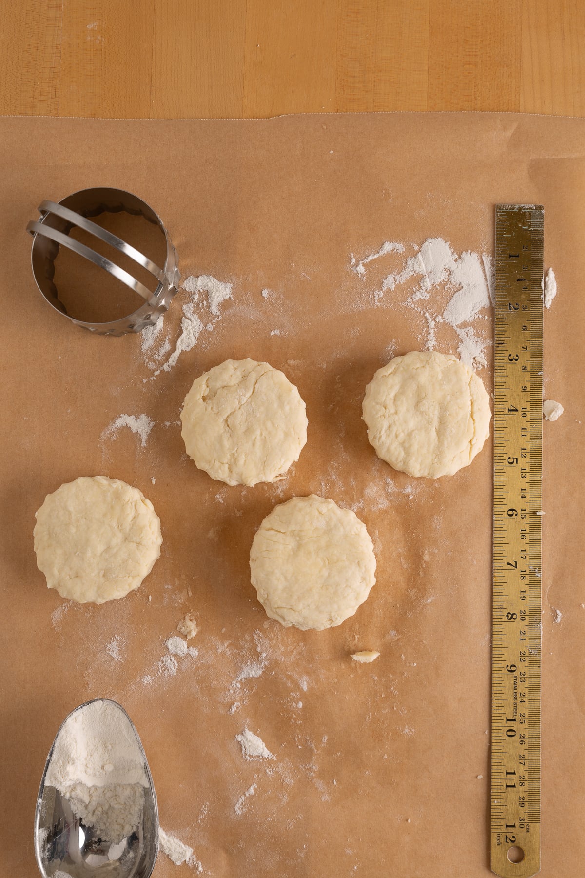 Four unbaked biscuit rounds on floured parchment paper, with a biscuit cutter, metal ruler, and flour scoop arranged nearby.