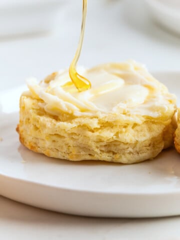 A biscuit with melted butter on a white plate, being drizzled with honey—freshly made air fryer biscuits for a warm and delicious treat.