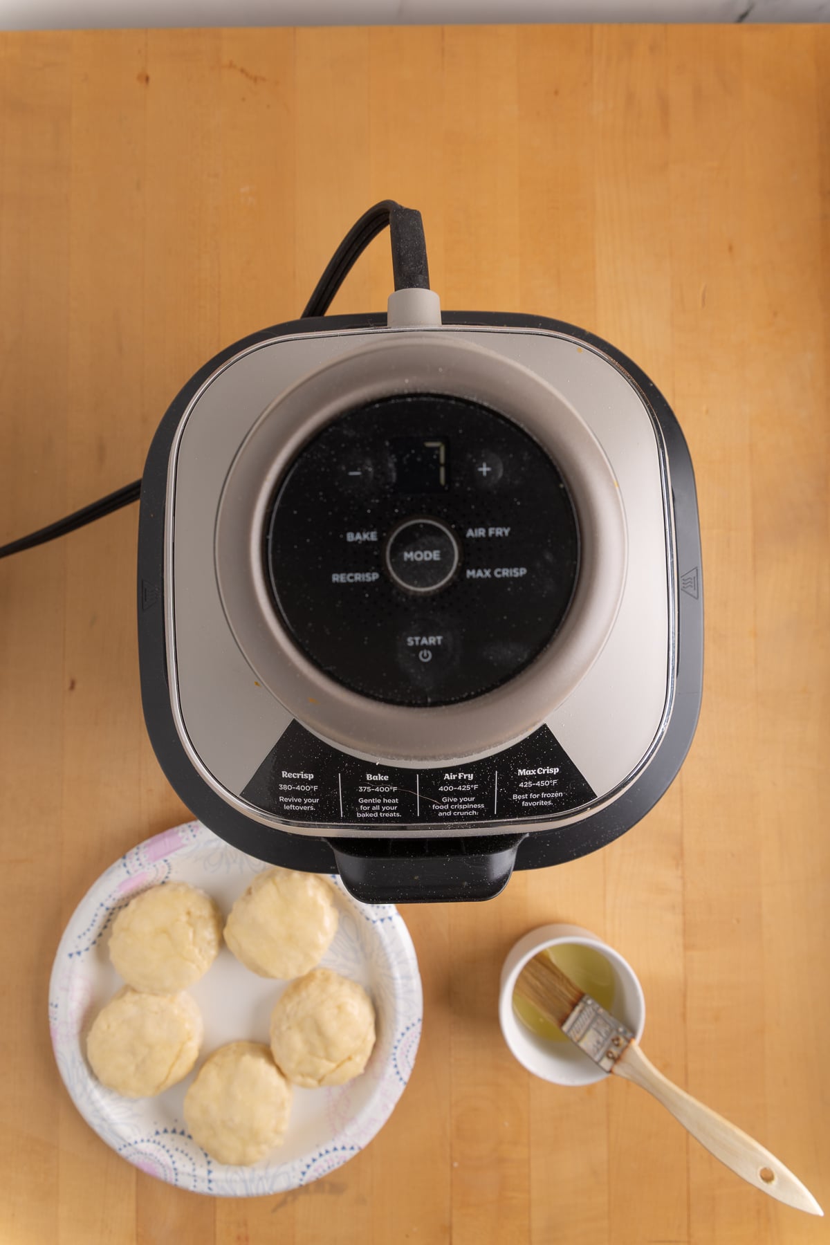 An air fryer sits on a wooden surface next to a paper plate with raw biscuit dough and a small bowl of melted butter with a brush.