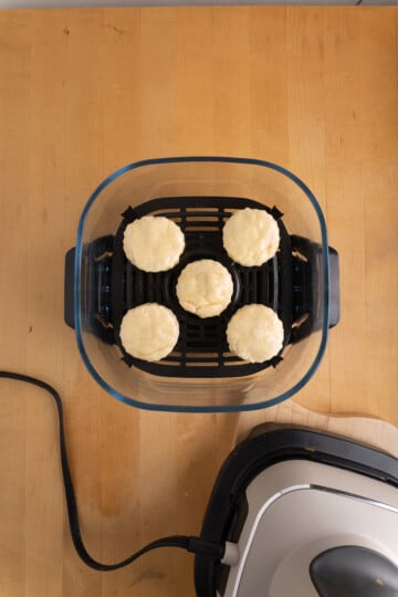 Six unbaked biscuits are arranged in a single layer inside an air fryer basket on a wooden surface.