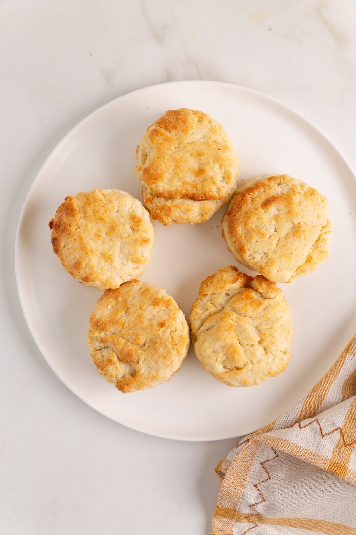 Five golden-brown biscuits are arranged in a circle on a white plate, with part of a beige and white cloth napkin visible nearby.