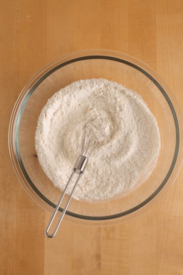 A glass bowl filled with flour and a metal whisk resting inside, placed on a wooden surface.