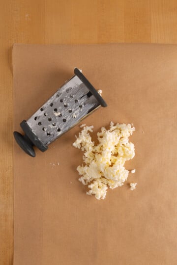 A box grater lies on brown parchment paper next to a small pile of grated butter on a wooden surface.
