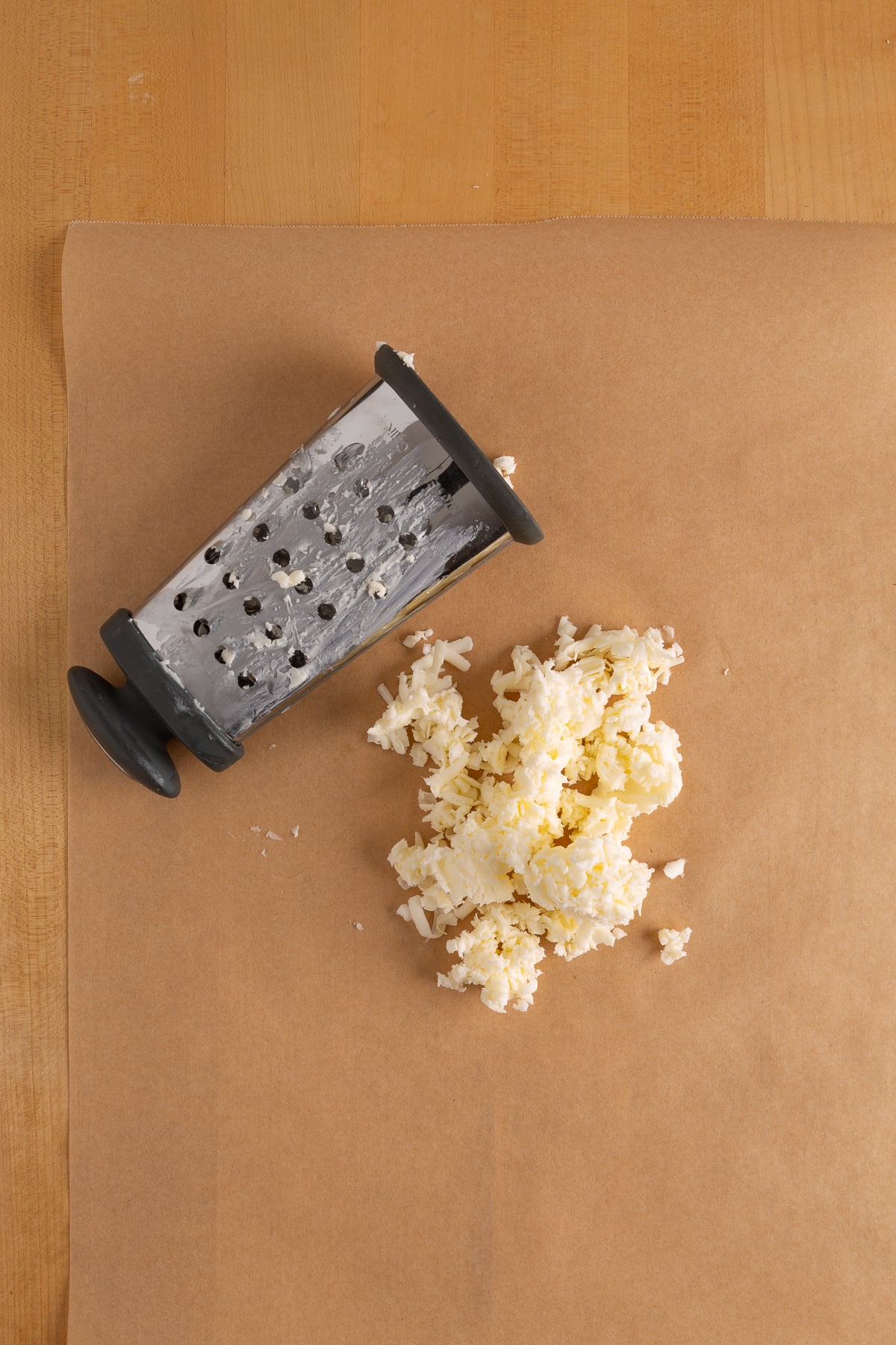 A box grater lies on brown parchment paper next to a small pile of grated butter on a wooden surface.