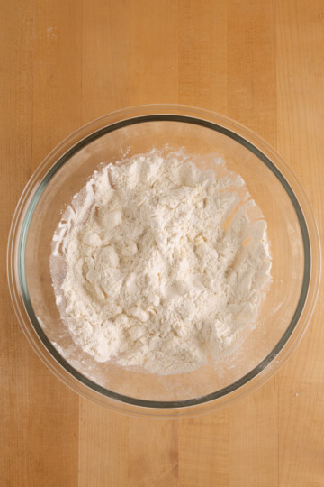 A clear glass bowl filled with flour sits on a wooden surface.