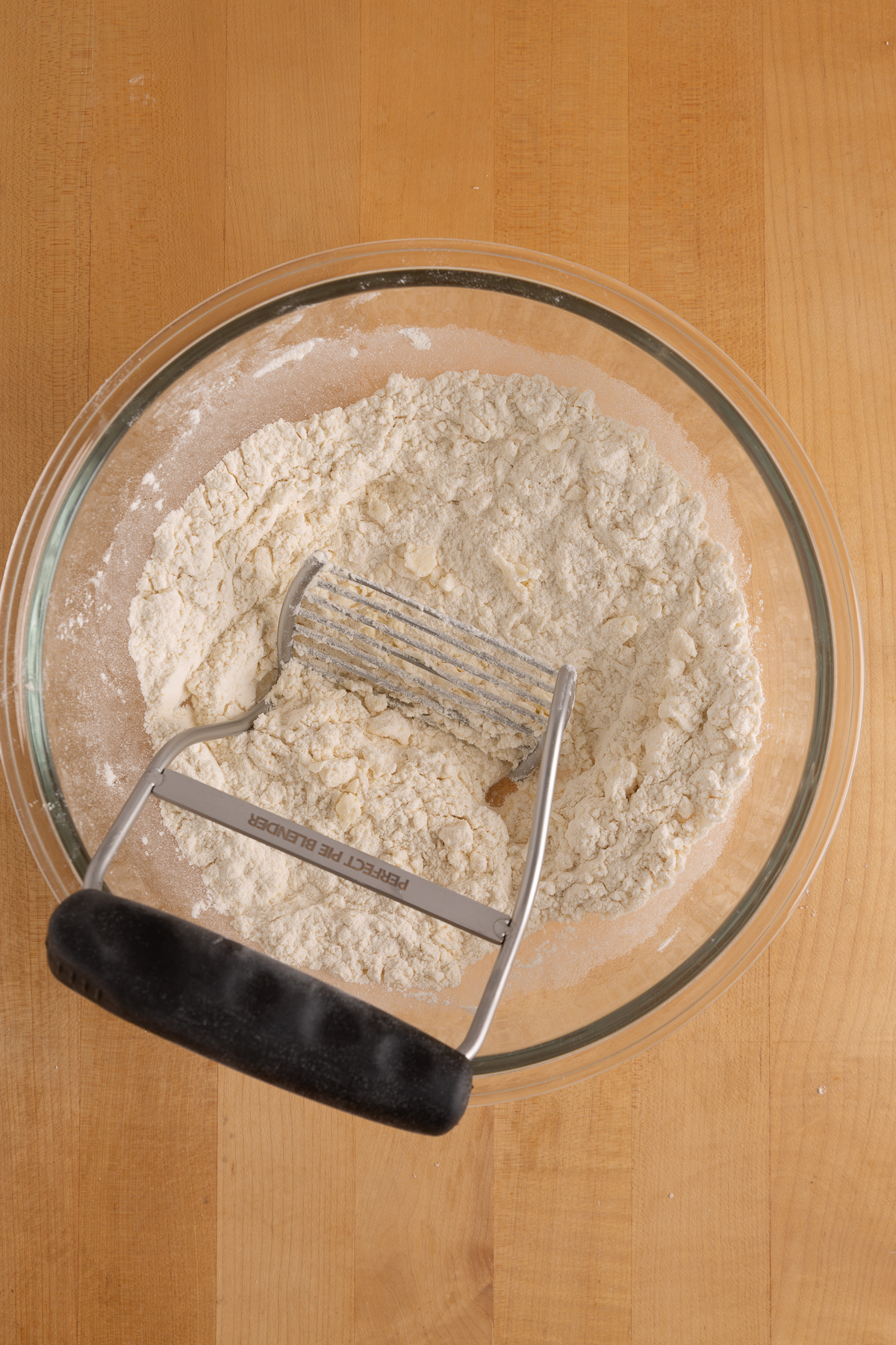 A glass bowl with flour mixture and a pastry blender rests on a wooden surface.