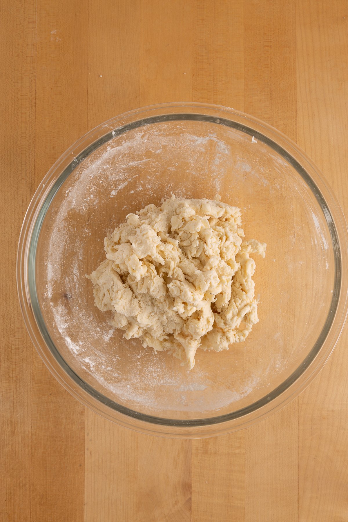 A clear glass bowl containing a rough, partially mixed dough sits on a light wooden surface.