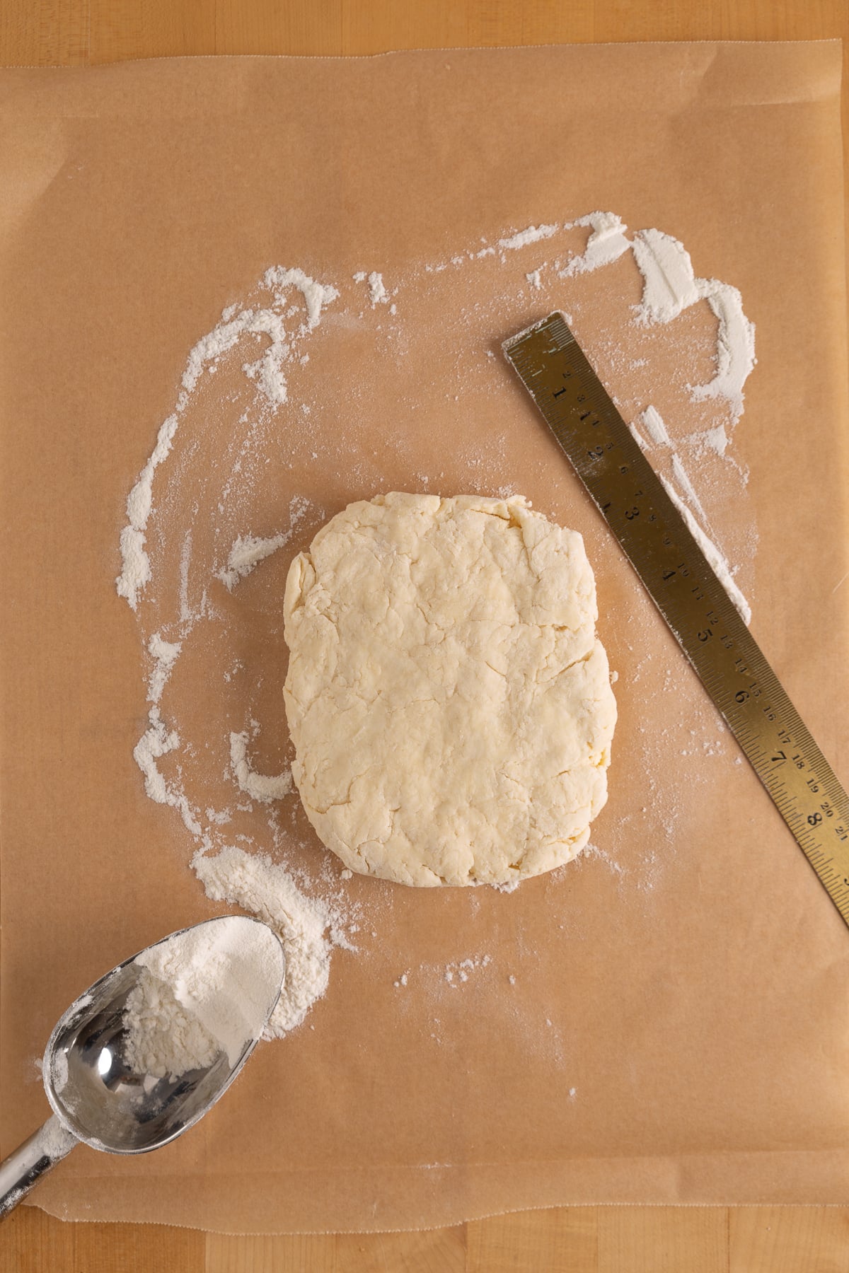 A square piece of dough sits on parchment paper with flour, a metal ruler, and a metal scoop holding more flour nearby.