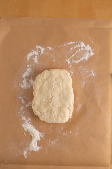 A square-shaped ball of dough rests on a sheet of brown parchment paper with scattered flour around it.