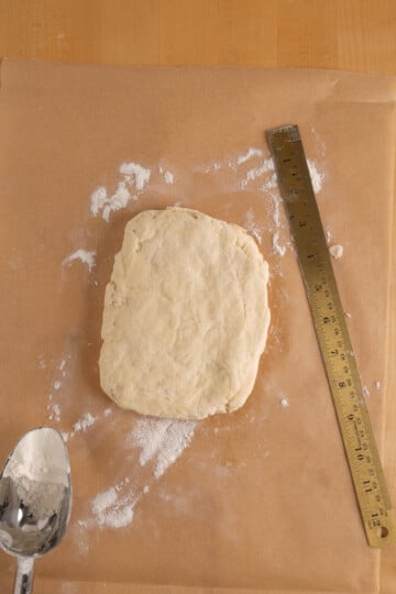 A square piece of dough rests on parchment paper beside a metal ruler and a floured spoon on a wooden surface.