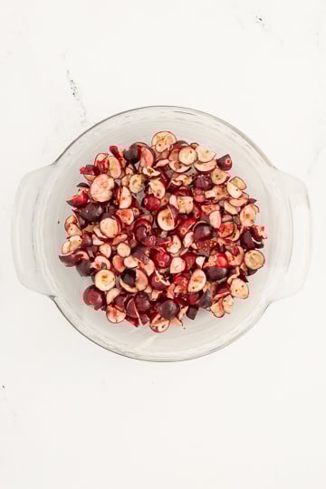 A glass bowl filled with halved and pitted cherries on a white surface.