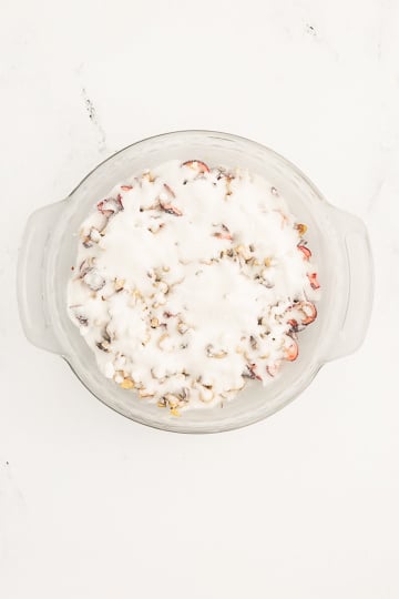 A round glass baking dish filled with chopped fruit and nuts, topped with a layer of white batter or cream, on a white surface.
