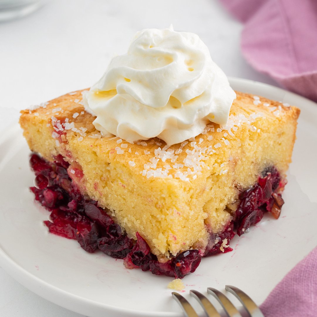 A slice of Nantucket cranberry pie topped with a swirl of whipped cream sits on a white plate next to a fork.