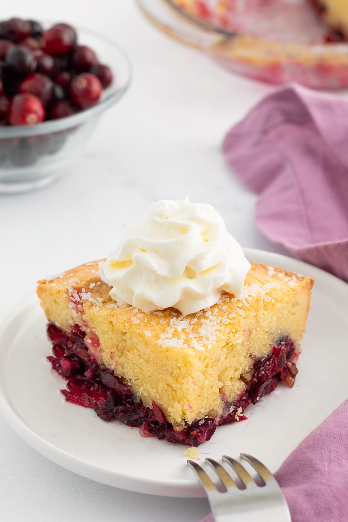 A slice of Nantucket cranberry pie topped with whipped cream on a white plate, with a fork, purple napkin, and a bowl of berries in the background.