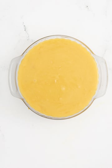 A round pie dish filled with an unbaked, smooth yellow batter on a white background.