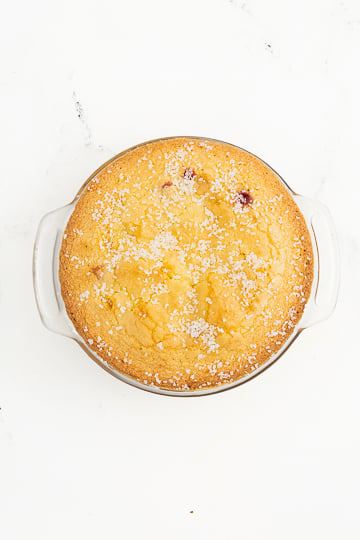 A round baked dessert in a white dish, topped with granulated sugar, on a white marble surface.
