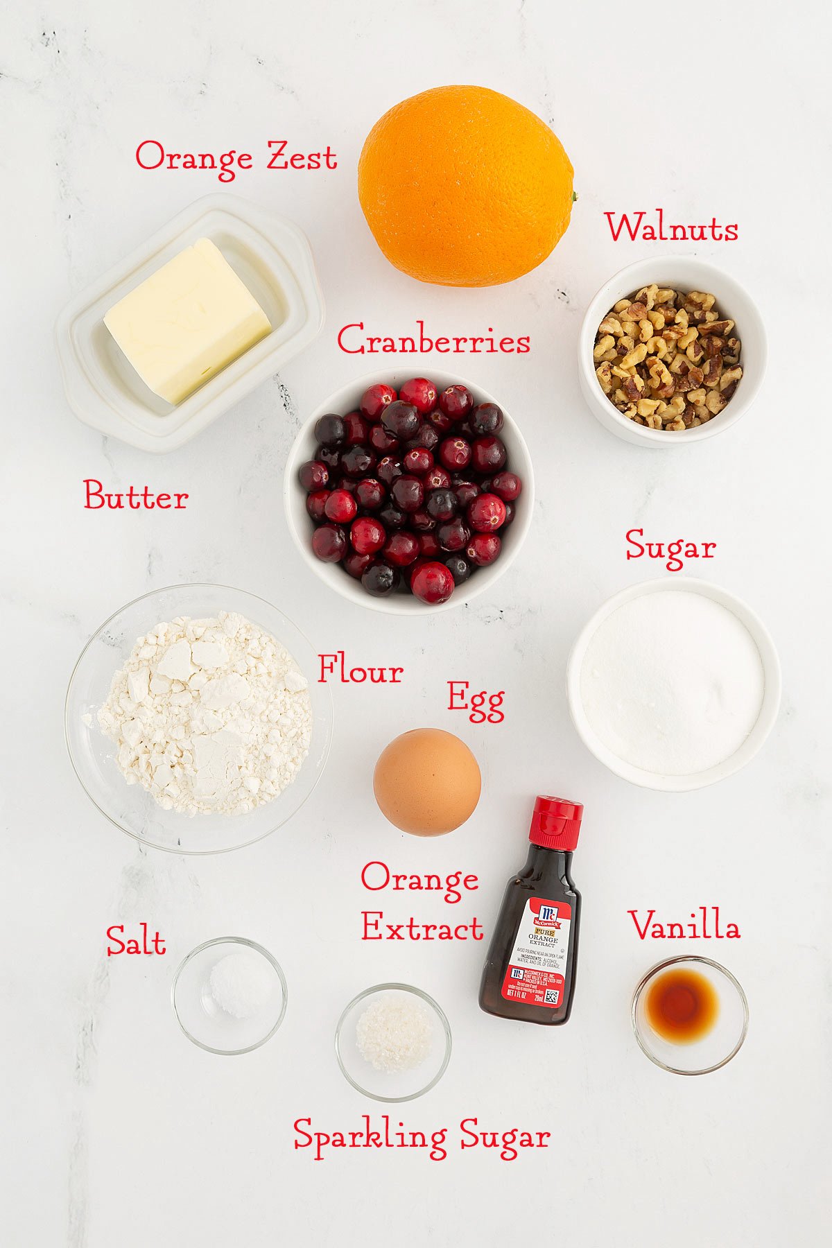 Top-down view of baking ingredients labeled: orange zest, walnuts, butter, cranberries, sugar, flour, egg, orange extract, vanilla, salt, and sparkling sugar on a white surface.