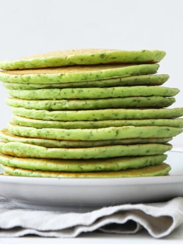 A stack of green pancakes is arranged on a white plate, which rests on a folded cloth napkin.
