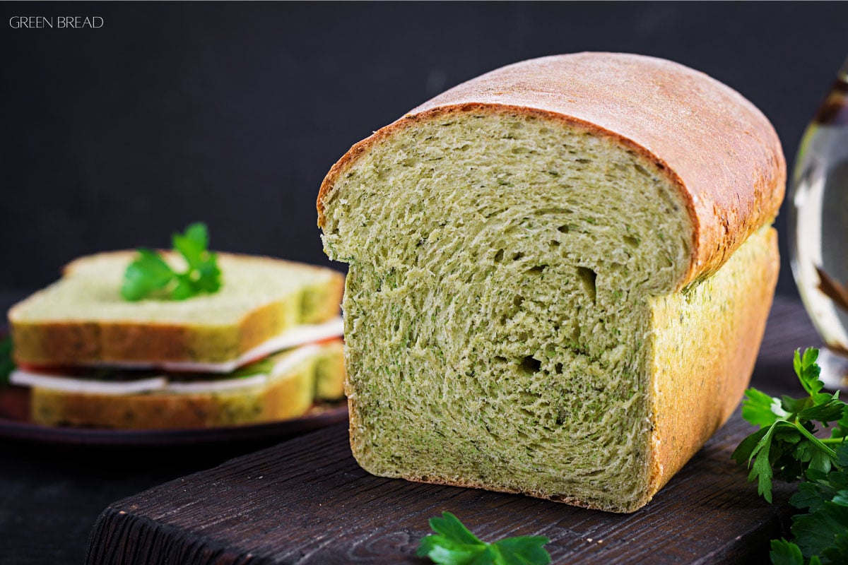 A loaf of green bread sits on a wooden surface, with sandwich slices and parsley in the background.