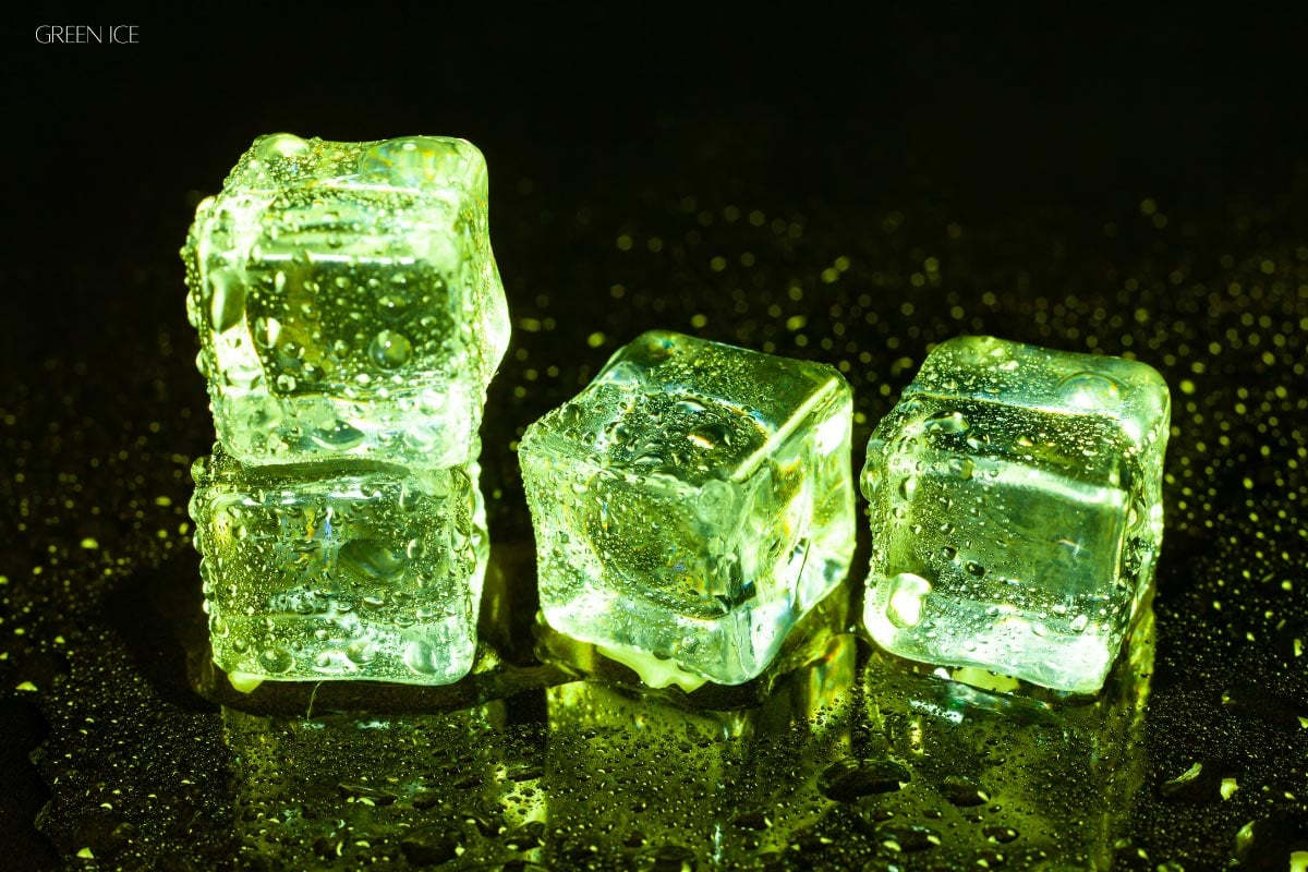 Four ice cubes illuminated with green light, with water droplets on their surfaces and scattered across a dark, reflective surface.